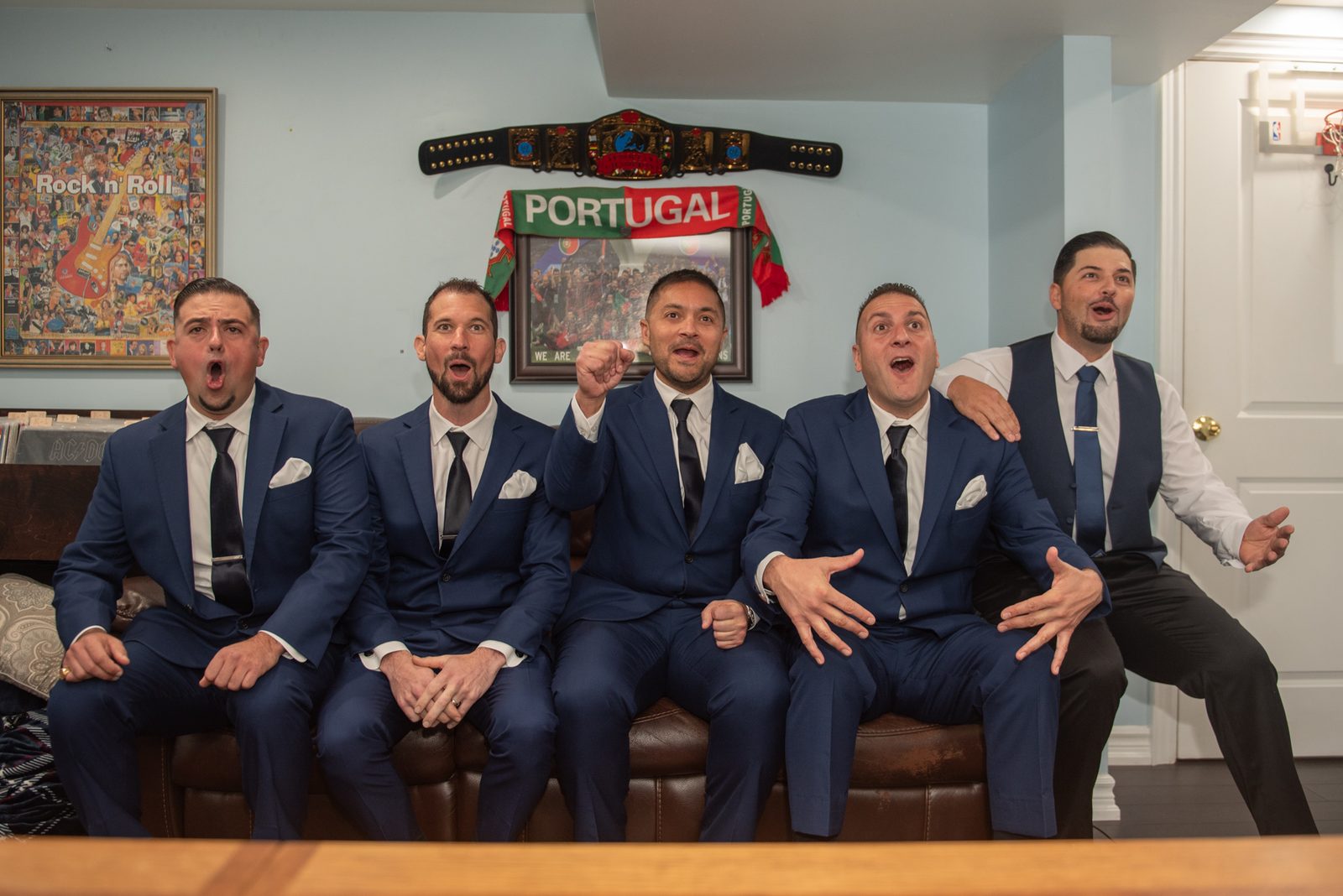 Five men in navy suits cheer exuberantly in a room decorated with Portugal memorabilia and a Rock n Roll poster.
