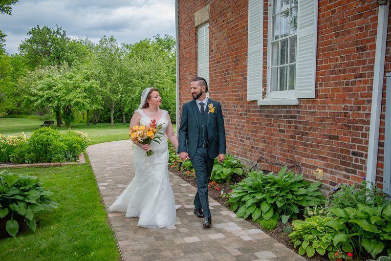 A bride in a veil and a groom in a classic suit walk hand in hand along a brick garden path beside a red brick wall, smiling.