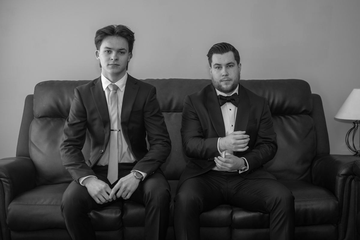Two formally dressed men sit side-by-side on a leather couch, looking at the camera with serious expressions.