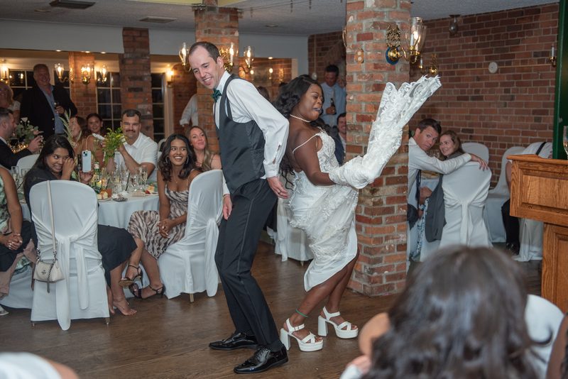 Bride and groom dance energetically in a warmly lit room with exposed brick walls, drawing laughter from guests.