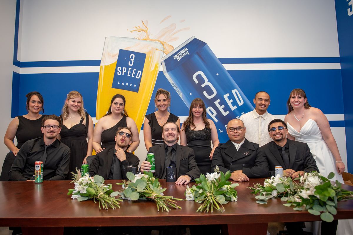 Formally dressed people pose around a table with white flowers and beer cans, behind a large promotional backdrop.