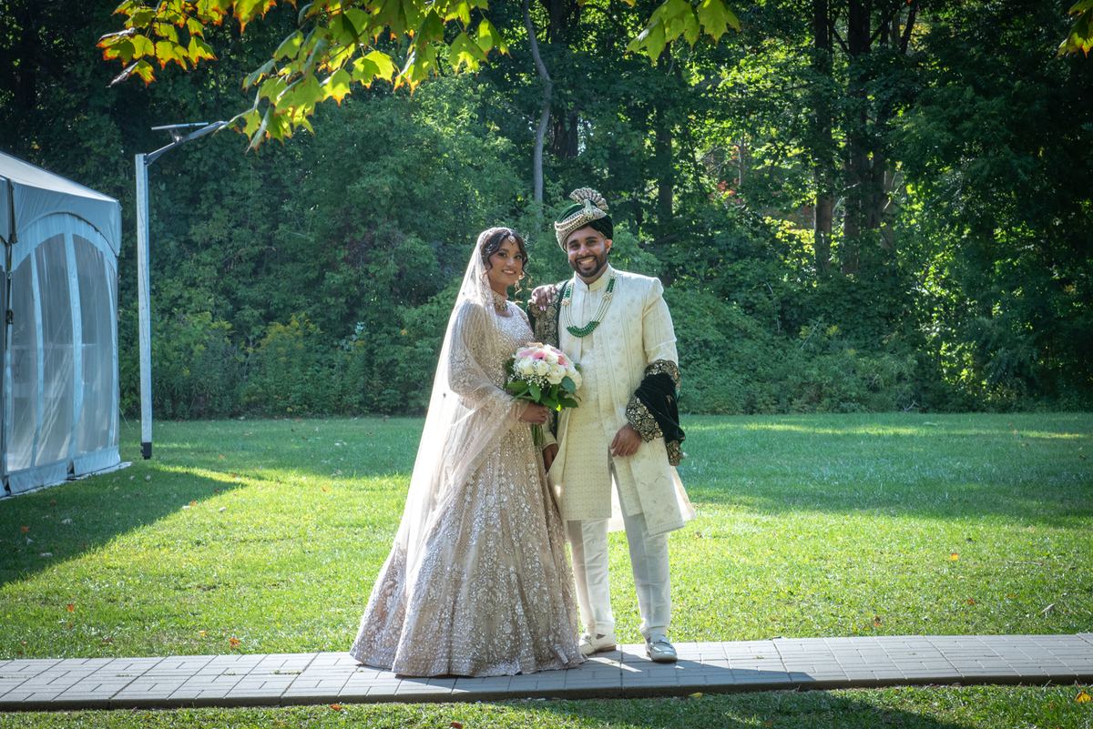 A smiling bride in a cream lehenga and groom in a matching sherwani stand on a garden path, sunlight filtering through trees.