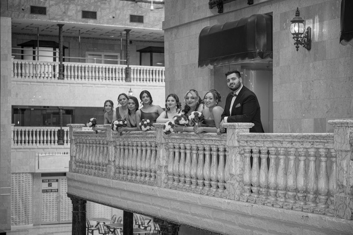Bride, groom, and bridesmaids lean casually on an ornate marble balcony inside a grand building, illuminated by soft light.
