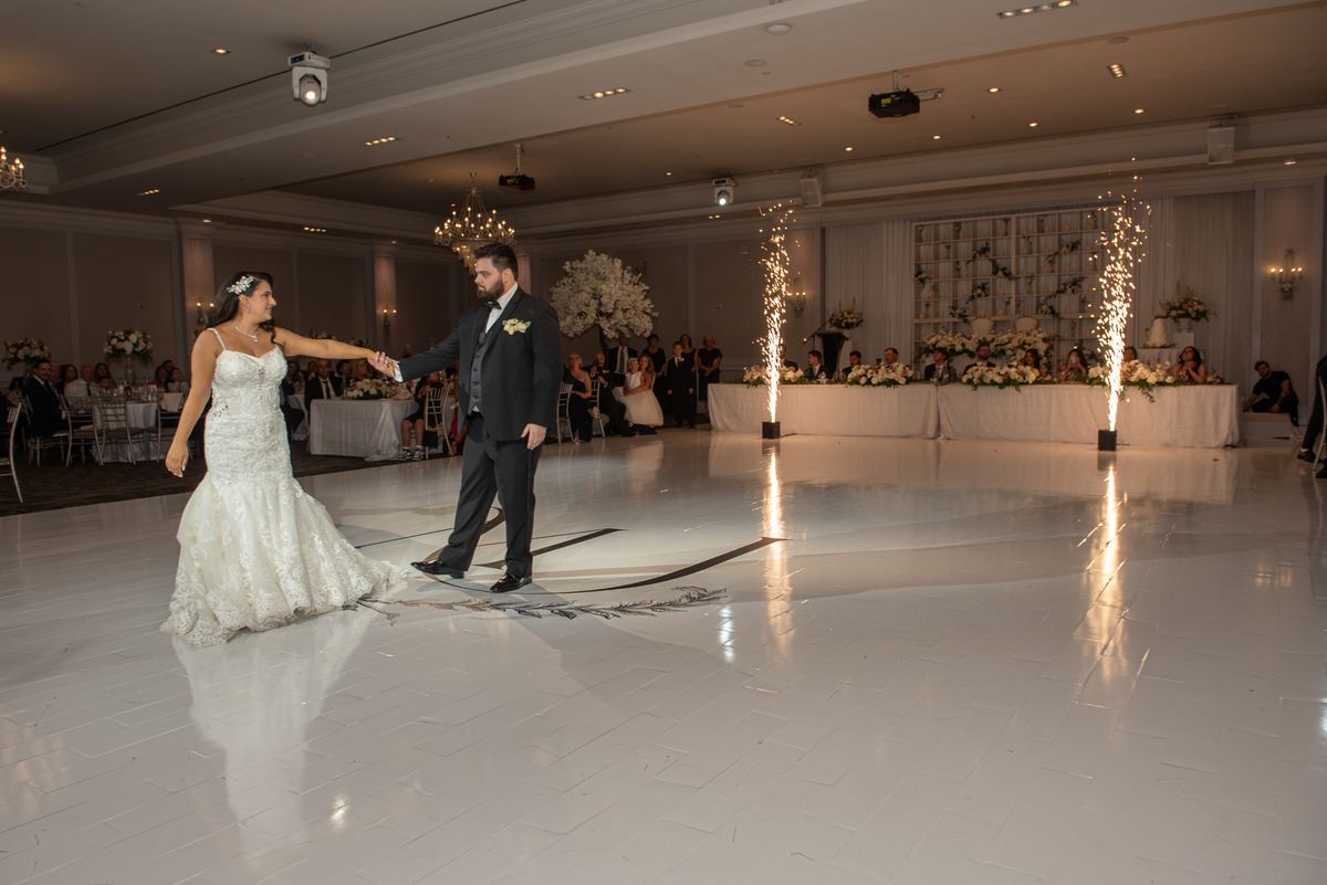 A bride and groom dance on a white ballroom floor surrounded by decorated tables and sparkler columns under warm lighting.