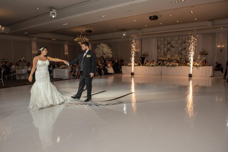 A bride and groom dance on a white ballroom floor surrounded by decorated tables and sparkler columns under warm lighting.