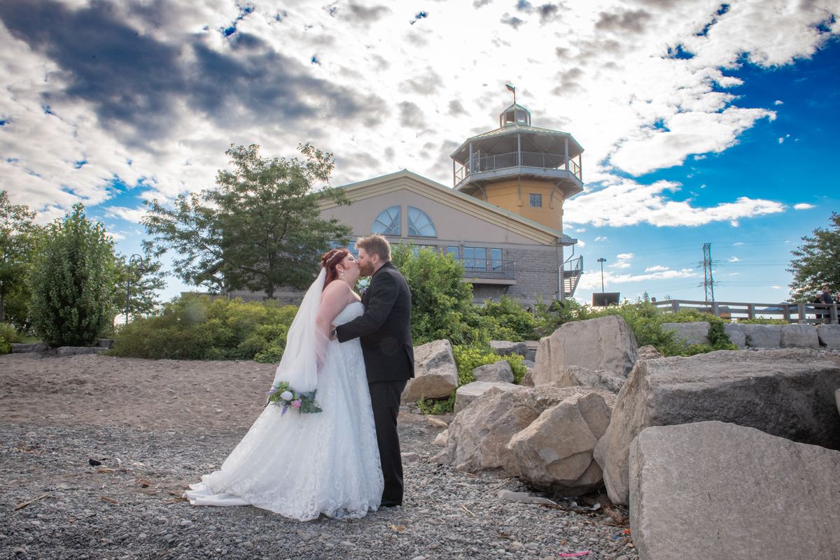 A bride and groom kiss beside a lighthouse-style building on a gravel path surrounded by rocks and greenery.