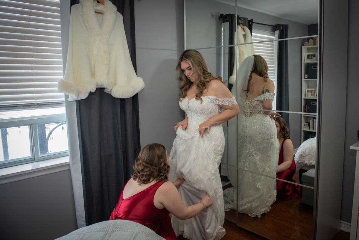 A bride adjusts her wedding gown in front of a mirror as a woman in red assists, with a white fur cape hanging nearby.