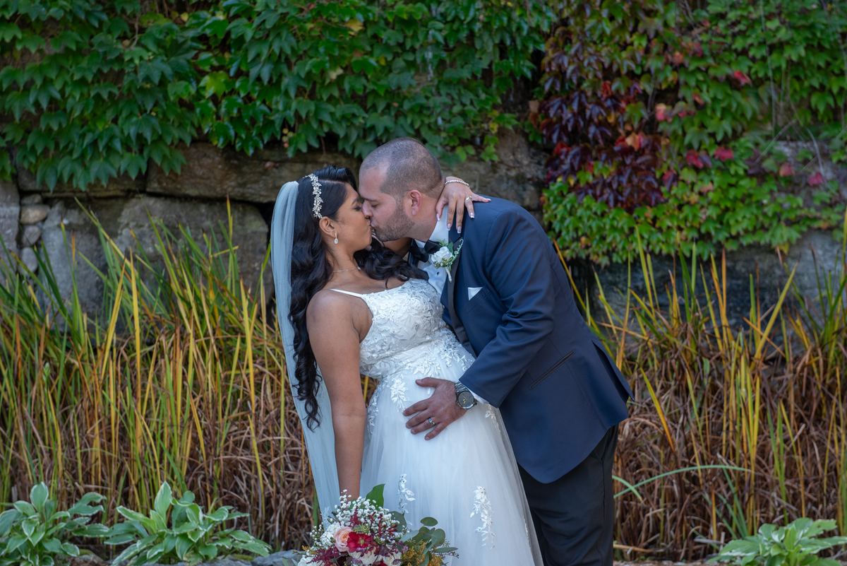 A bride and groom kiss outdoors surrounded by greenery and autumn leaves, bathed in soft natural light.