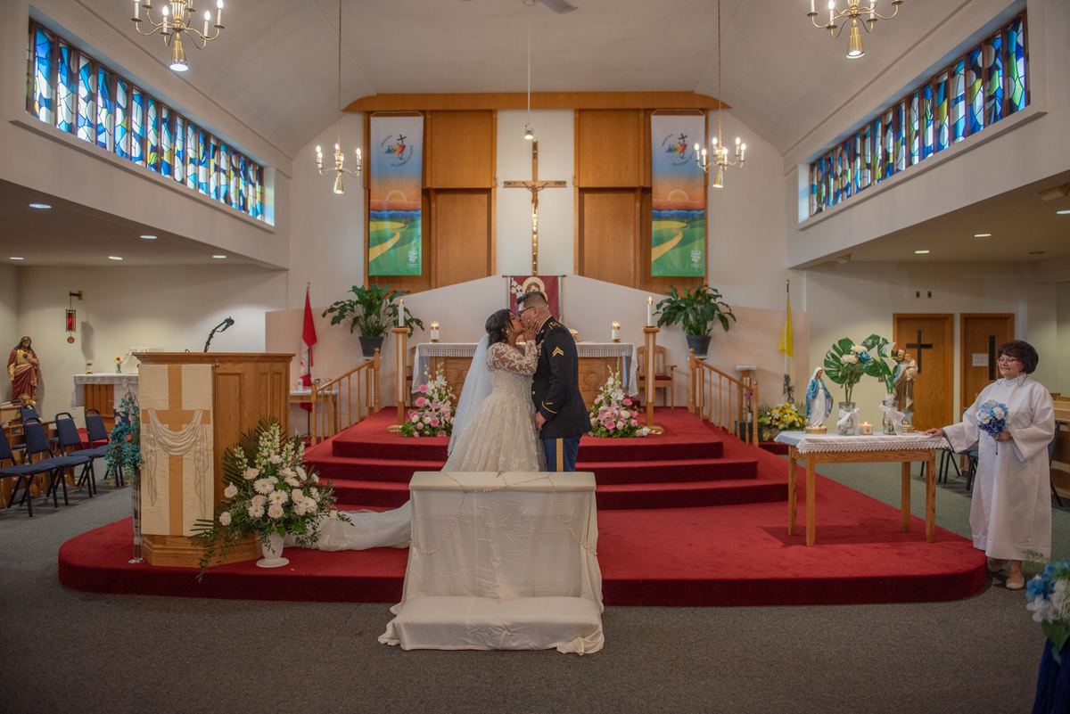 Bride and groom kiss at an altar surrounded by flowers and stained glass, with a woman holding a bouquet nearby.