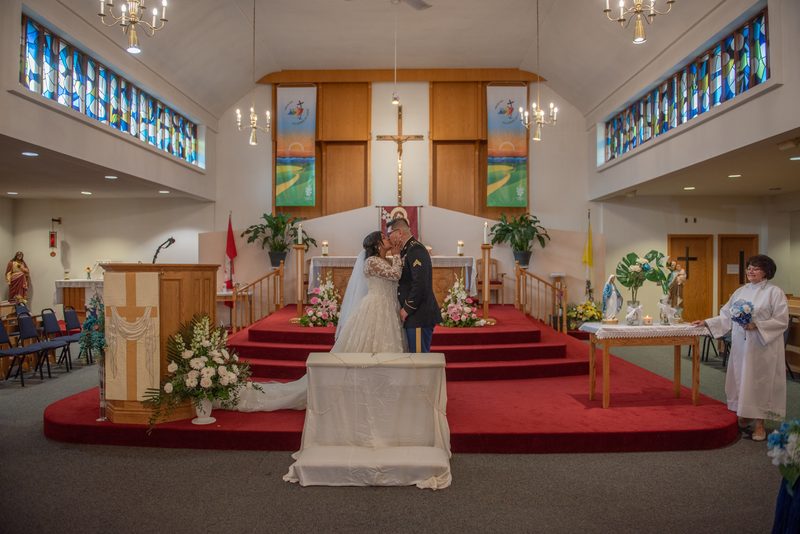 Bride and groom kiss at an altar surrounded by flowers and stained glass, with a woman holding a bouquet nearby.