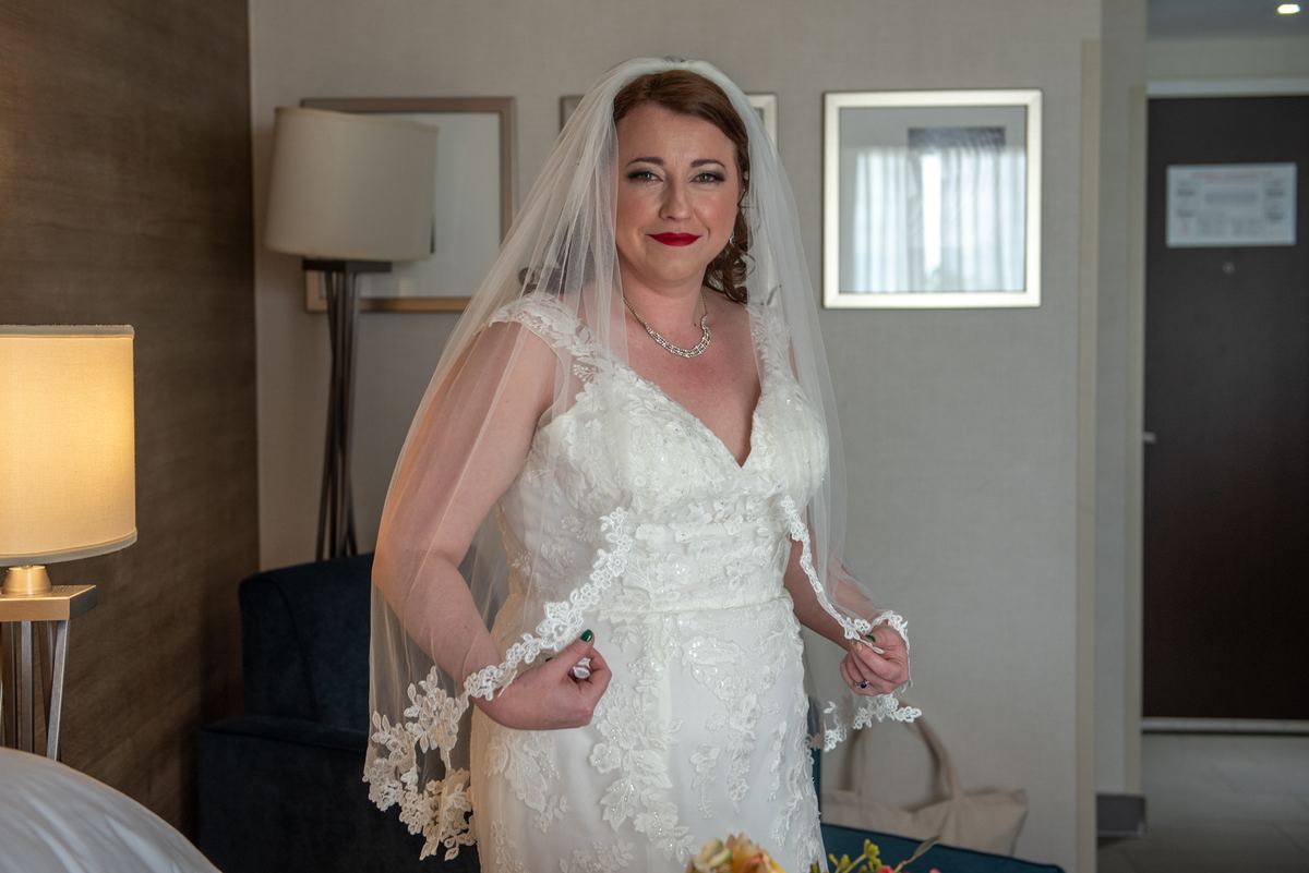 A bride in a hotel room, smiling and adjusting her lace-trimmed veil while holding the edge of her white wedding gown.