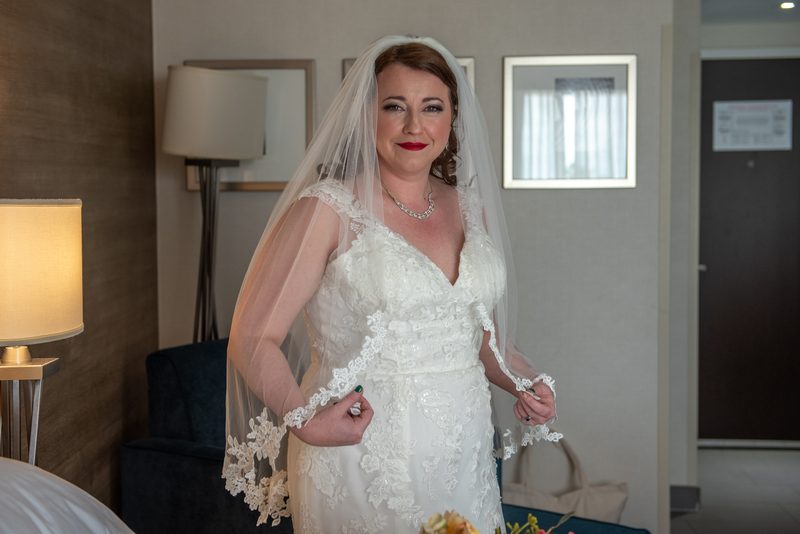 A bride in a hotel room, smiling and adjusting her lace-trimmed veil while holding the edge of her white wedding gown.