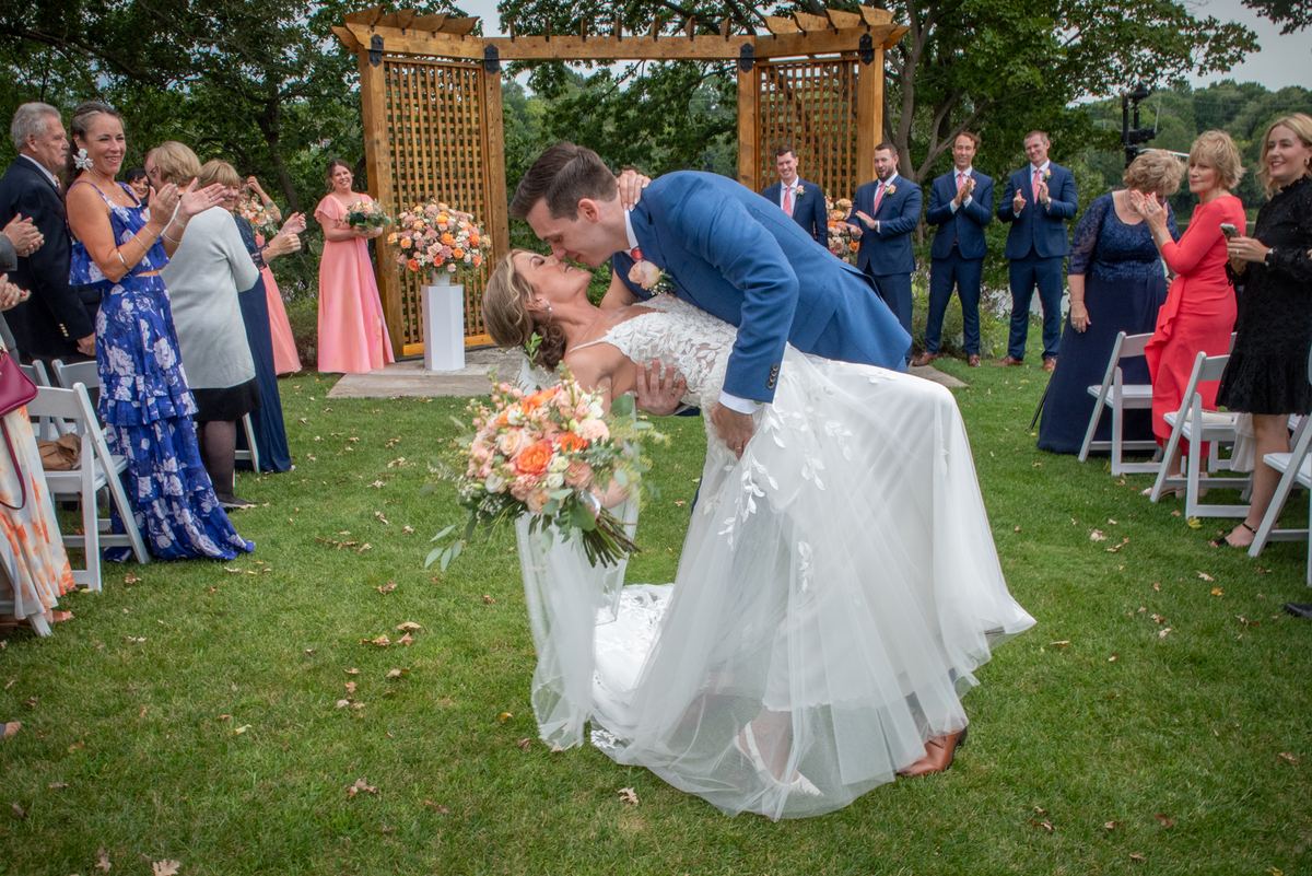Bride and groom kiss under wooden arbor surrounded by clapping guests in daylight, adorned with floral arrangements.