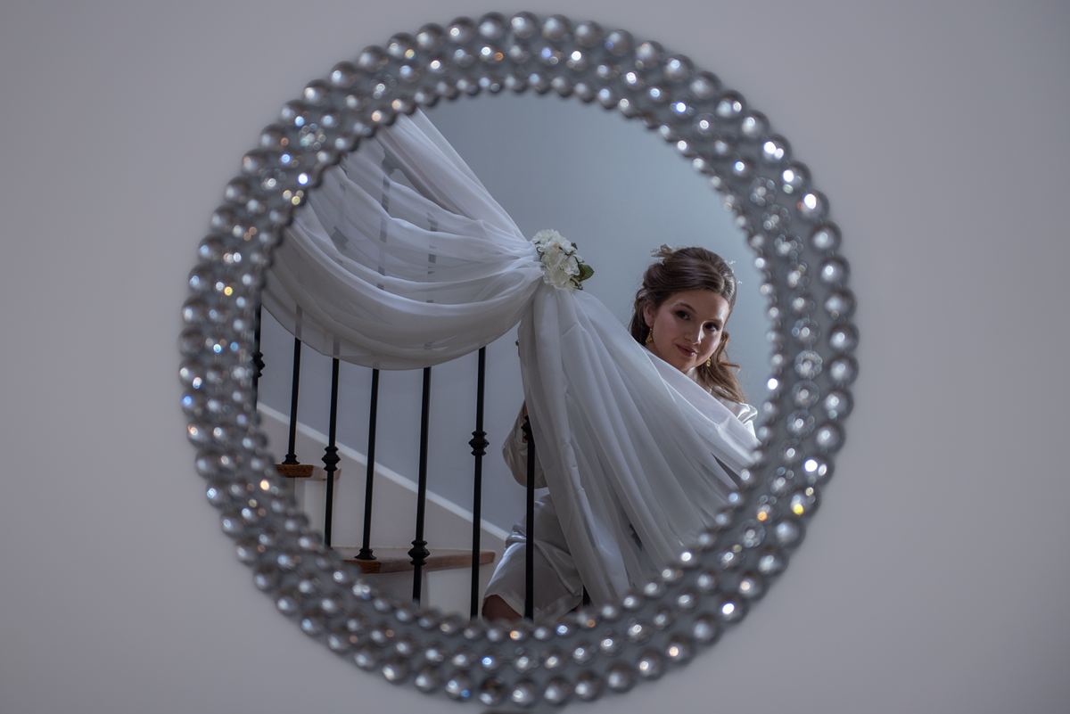 A bride, her veil cascading over a round pearl-encrusted mirror, stands introspectively on a staircase.