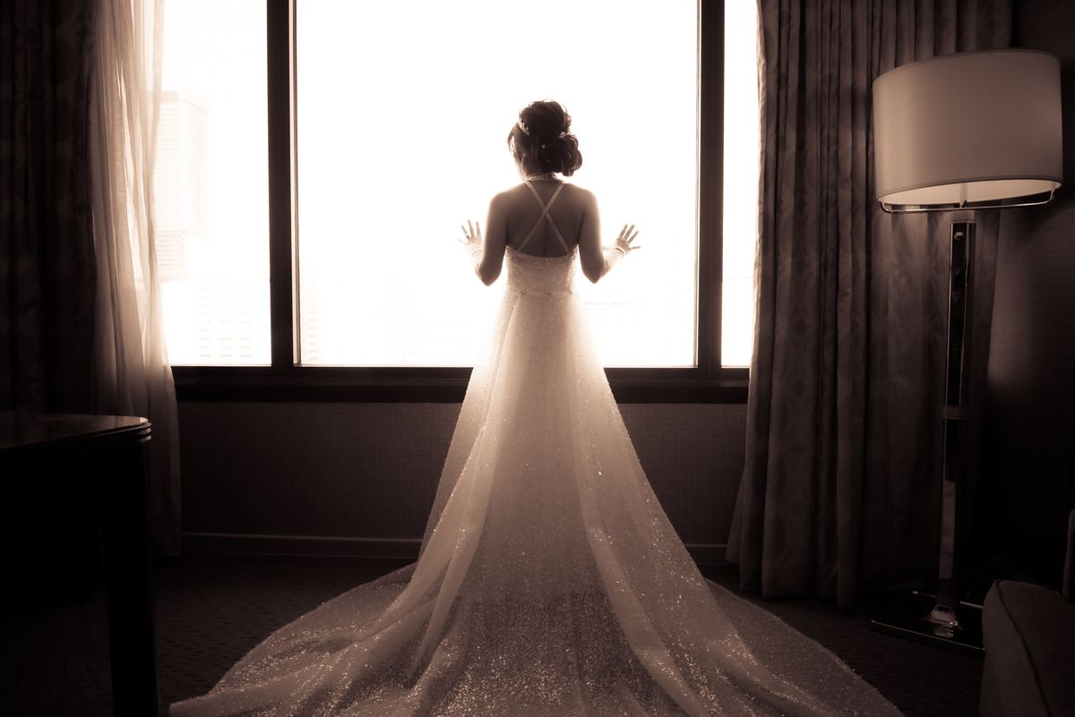 A bride in a sparkling gown stands by a bright window, her silhouette visible against the golden backlight of early evening.