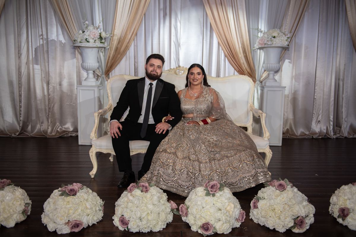 A bride in an ornate lehenga and groom in a tuxedo pose on a white settee, surrounded by floral arrangements and curtains.