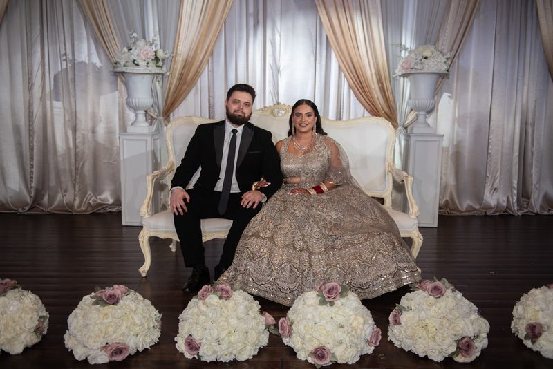 A bride in an ornate lehenga and groom in a tuxedo pose on a white settee, surrounded by floral arrangements and curtains.
