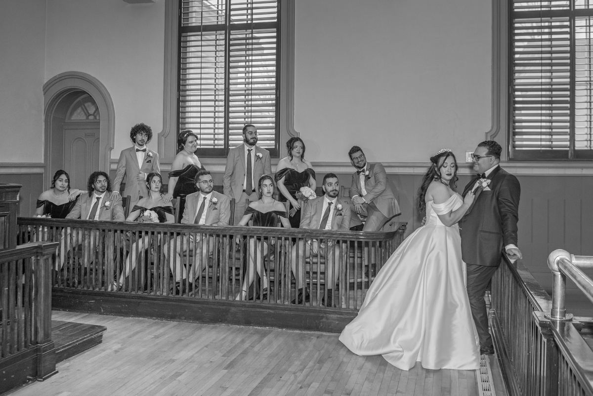Bride and groom stand on a platform in a historic courtroom, surrounded by their wedding party, smiling.