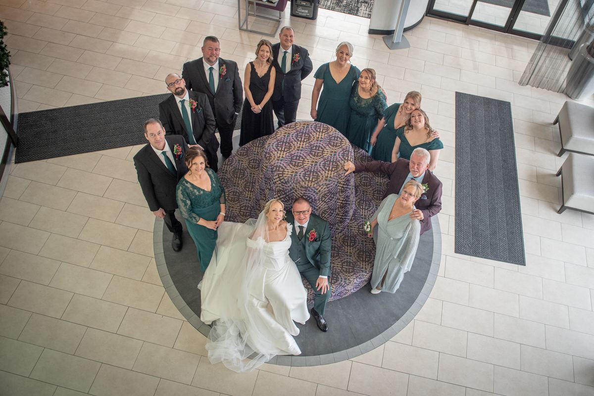 A bride and groom with their wedding party pose around an ottoman in a modern lobby, bathed in natural light.