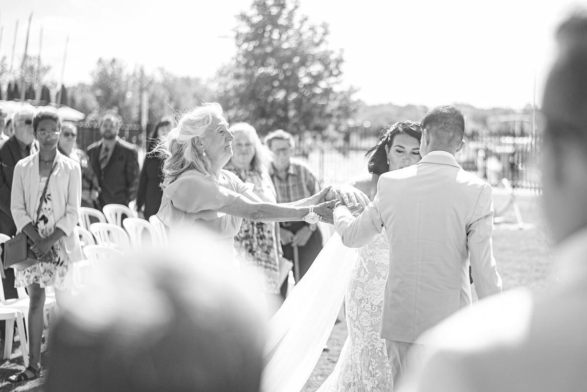 A bride and groom hold hands as an older woman guides them gently during an outdoor wedding ceremony.