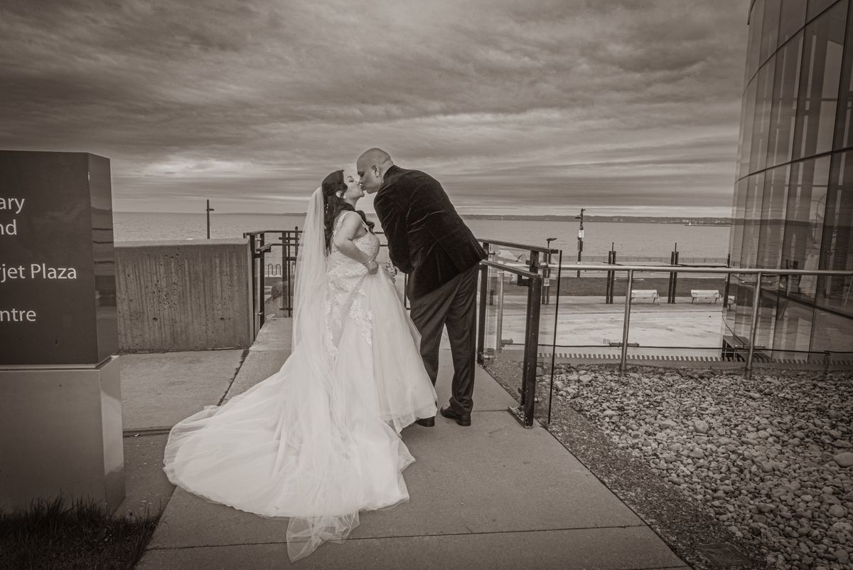A bride and groom kiss on a waterfront promenade, surrounded by modern architecture and an overcast sky.