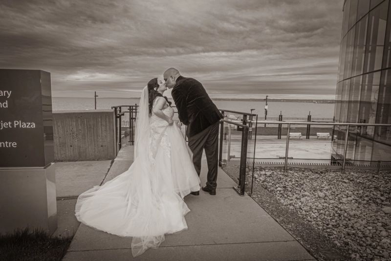 A bride and groom kiss on a waterfront promenade, surrounded by modern architecture and an overcast sky.