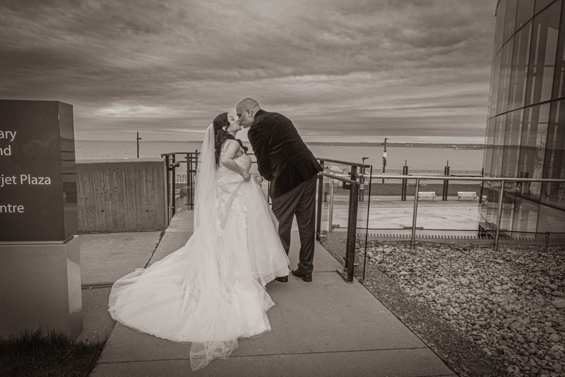 A bride and groom kiss on a waterfront promenade, surrounded by modern architecture and an overcast sky.