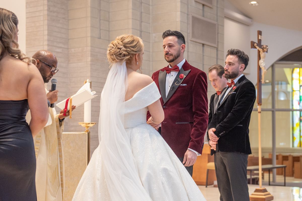 A bride in white and groom in maroon stand with officiant during church ceremony under stained glass light.