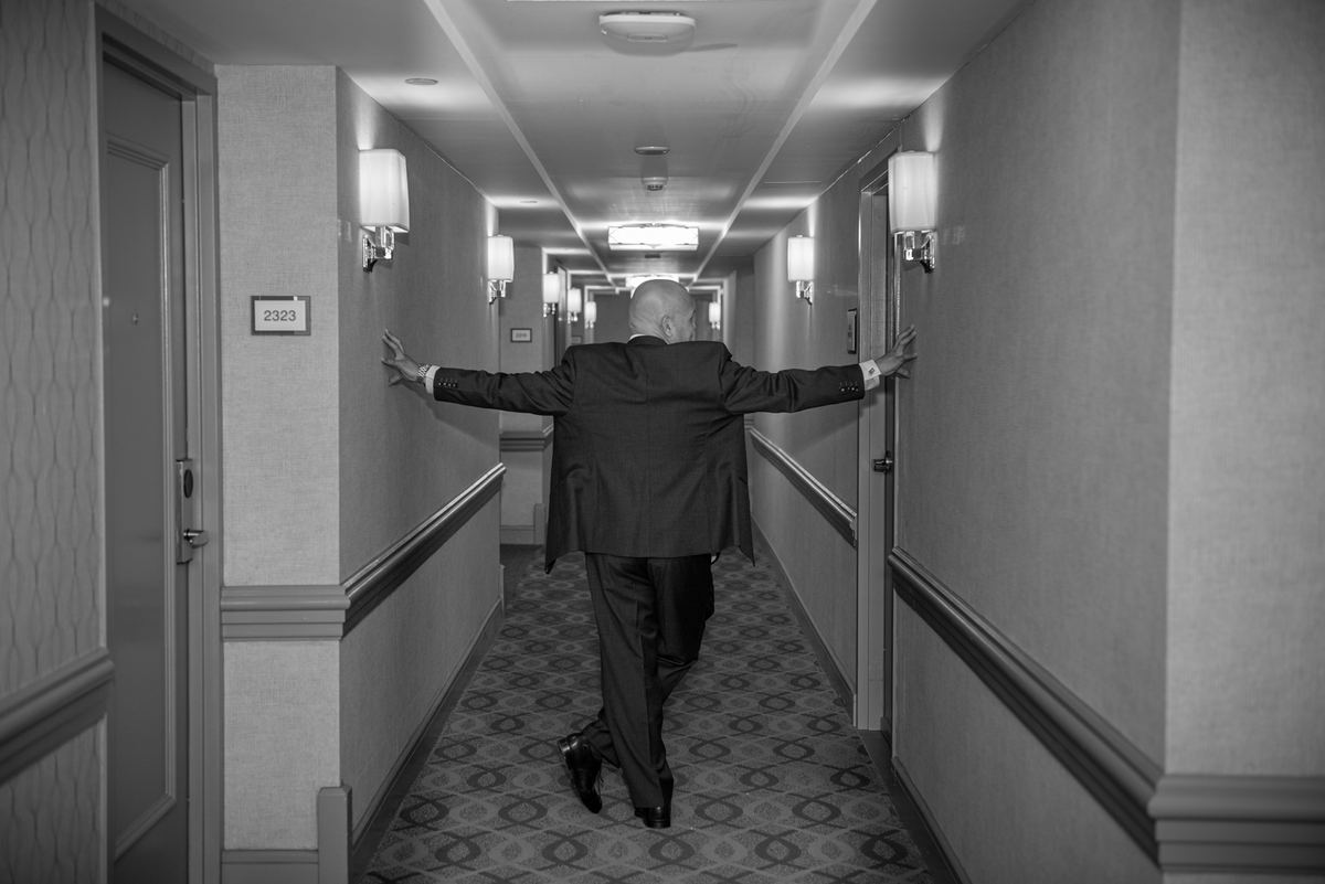 A man in a dark suit walks down a monochrome hotel corridor, touching the walls, in a moment of solitude.