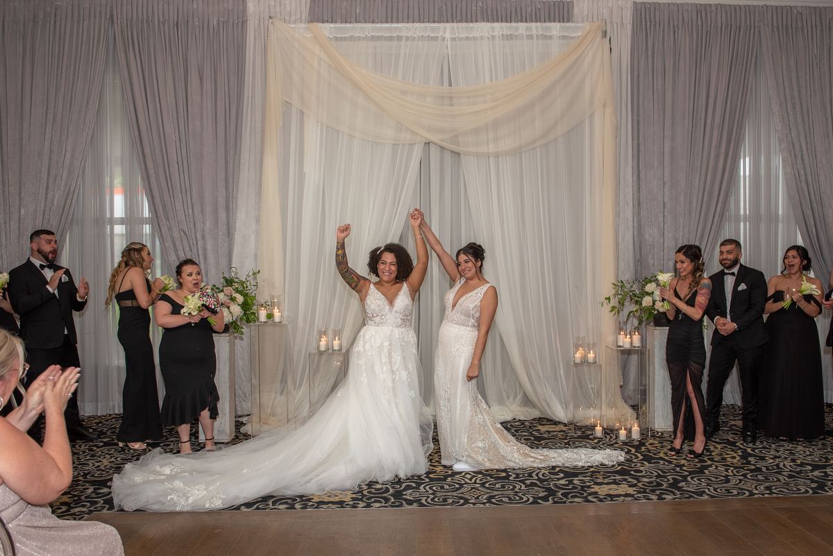 Two brides celebrate with raised arms at a wedding ceremony surrounded by smiling guests in formal attire.