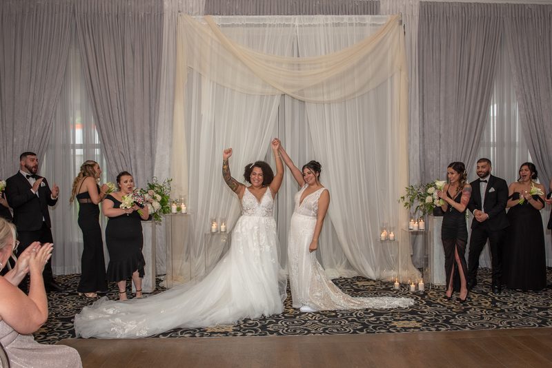 Two brides celebrate with raised arms at a wedding ceremony surrounded by smiling guests in formal attire.