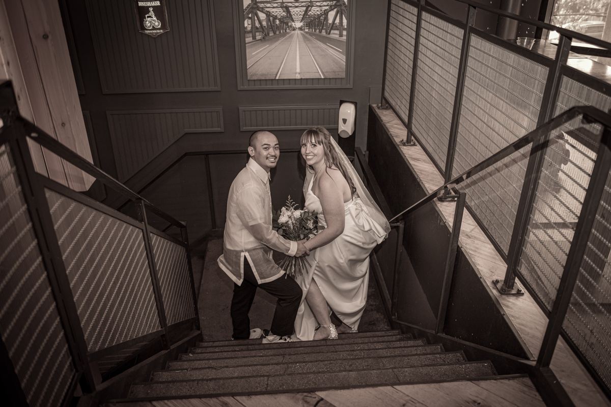 A newlywed couple poses joyfully at the top of a staircase, the groom presenting the bride with flowers.