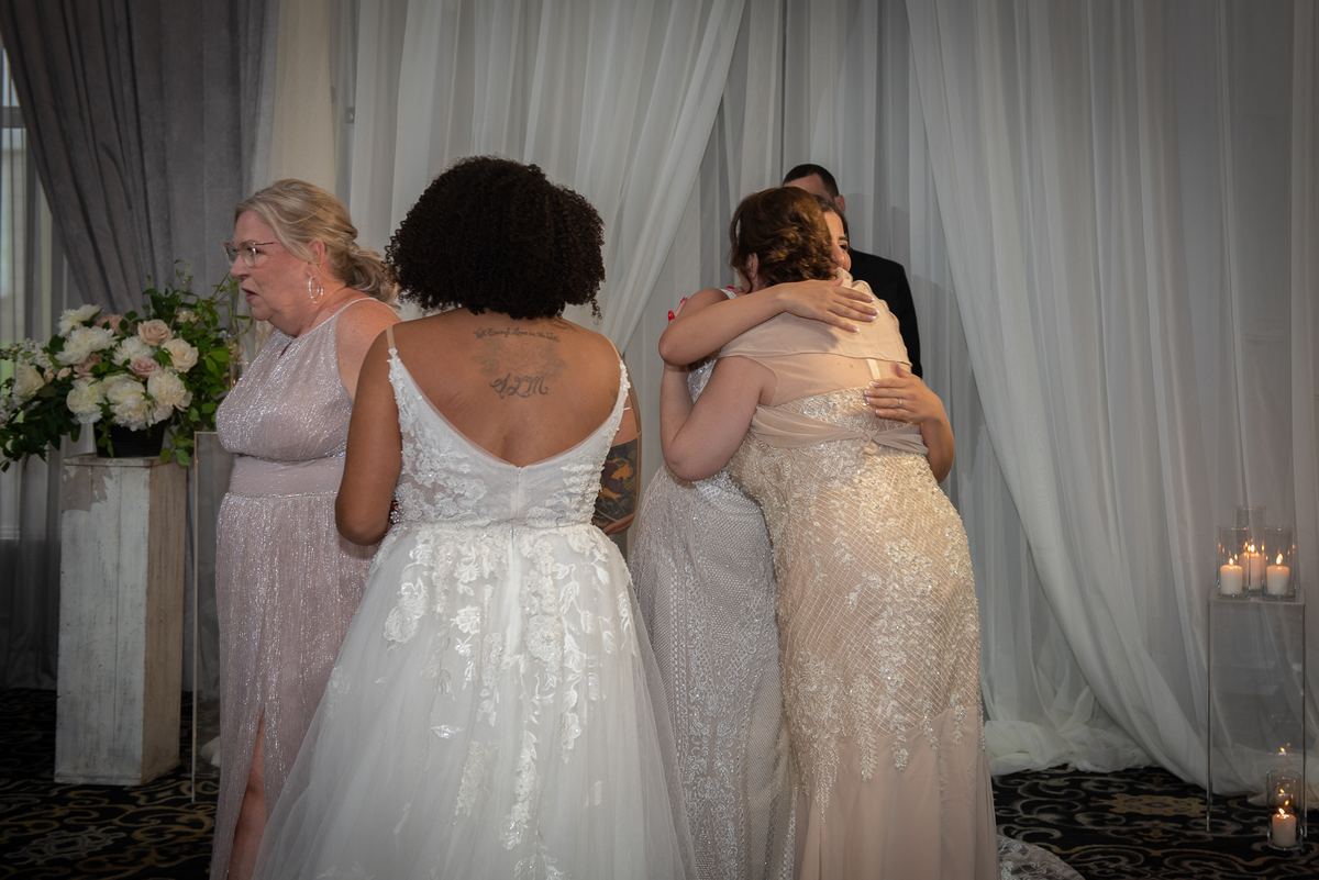Two brides embrace warmly during a wedding ceremony, with soft candlelight and white curtains creating a romantic atmosphere.