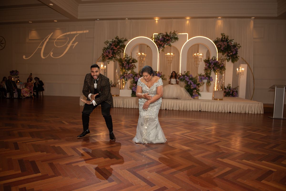 A bride and groom dance joyfully in a ballroom with an ornate floral arch, guests watching from tables.