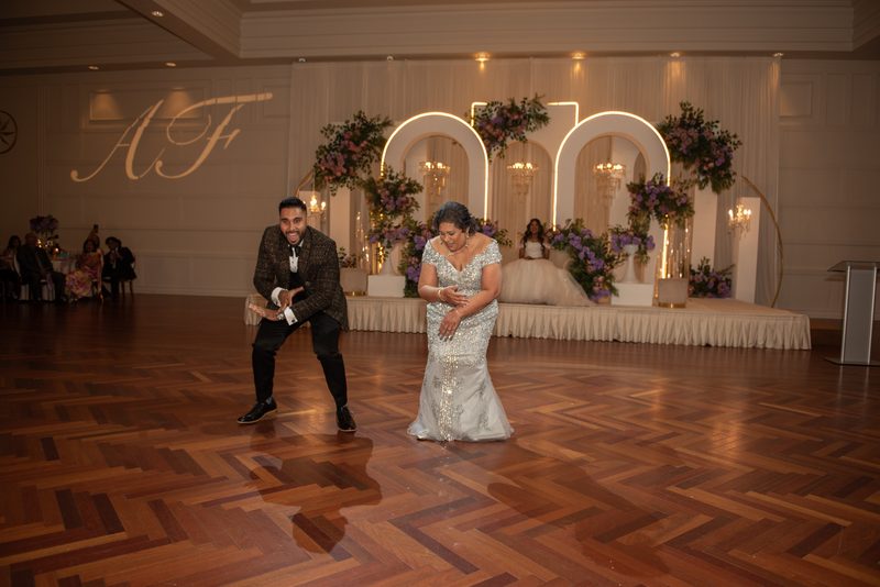 A bride and groom dance joyfully in a ballroom with an ornate floral arch, guests watching from tables.