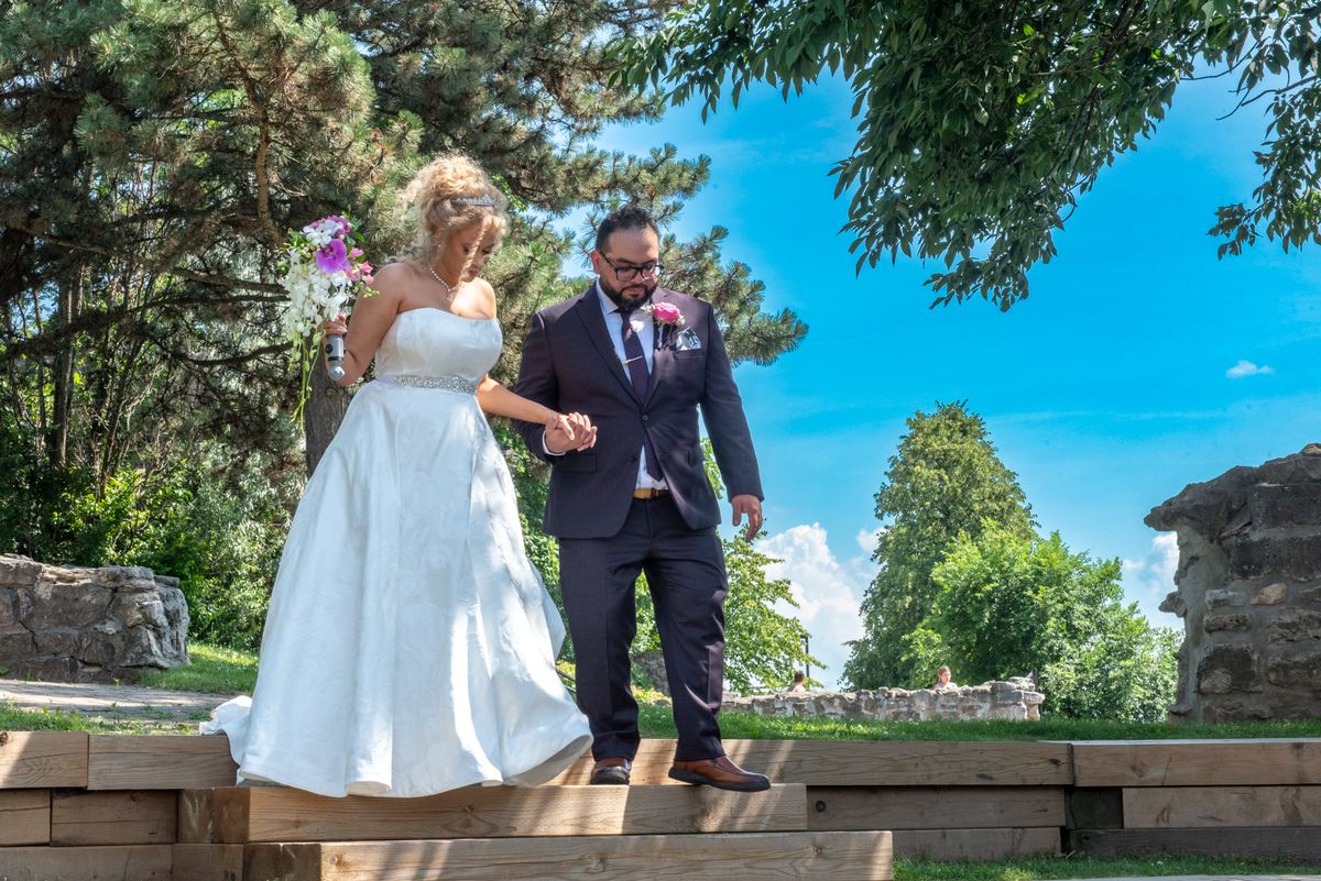 A bride in a white gown and groom in a dark suit walk hand-in-hand down wooden steps outdoors, surrounded by green trees.