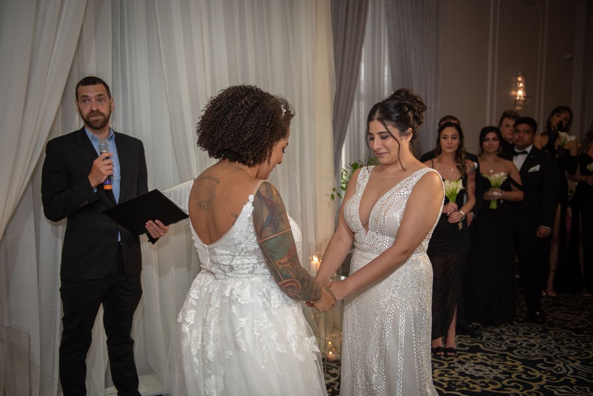 Two brides in formal attire hold hands during a ceremony, one with visible tattoos, under sheer curtains.