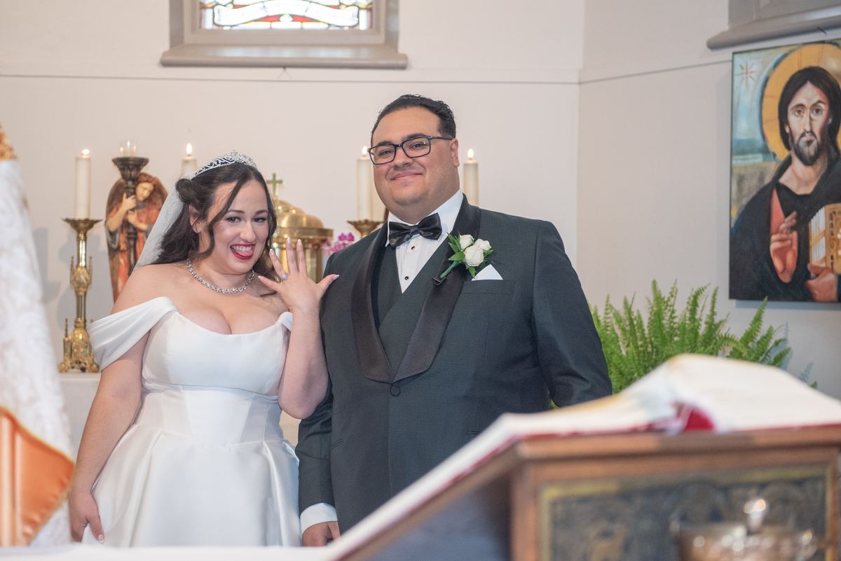A smiling bride and groom in wedding attire wave inside a church with candlelight and stained glass, celebrating their union.