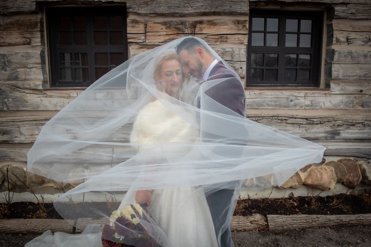 A bride and groom embrace in front of a rustic log cabin, their veiled faces close as the wind blows.