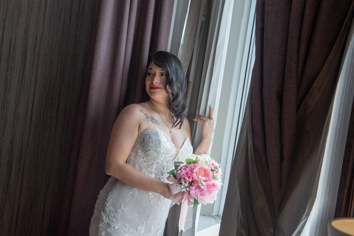 A bride in a lace gown holds a pink and white bouquet by a window, smiling softly at something off-camera.