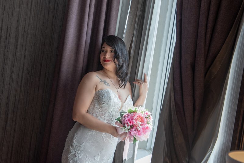 A bride in a lace gown holds a pink and white bouquet by a window, smiling softly at something off-camera.