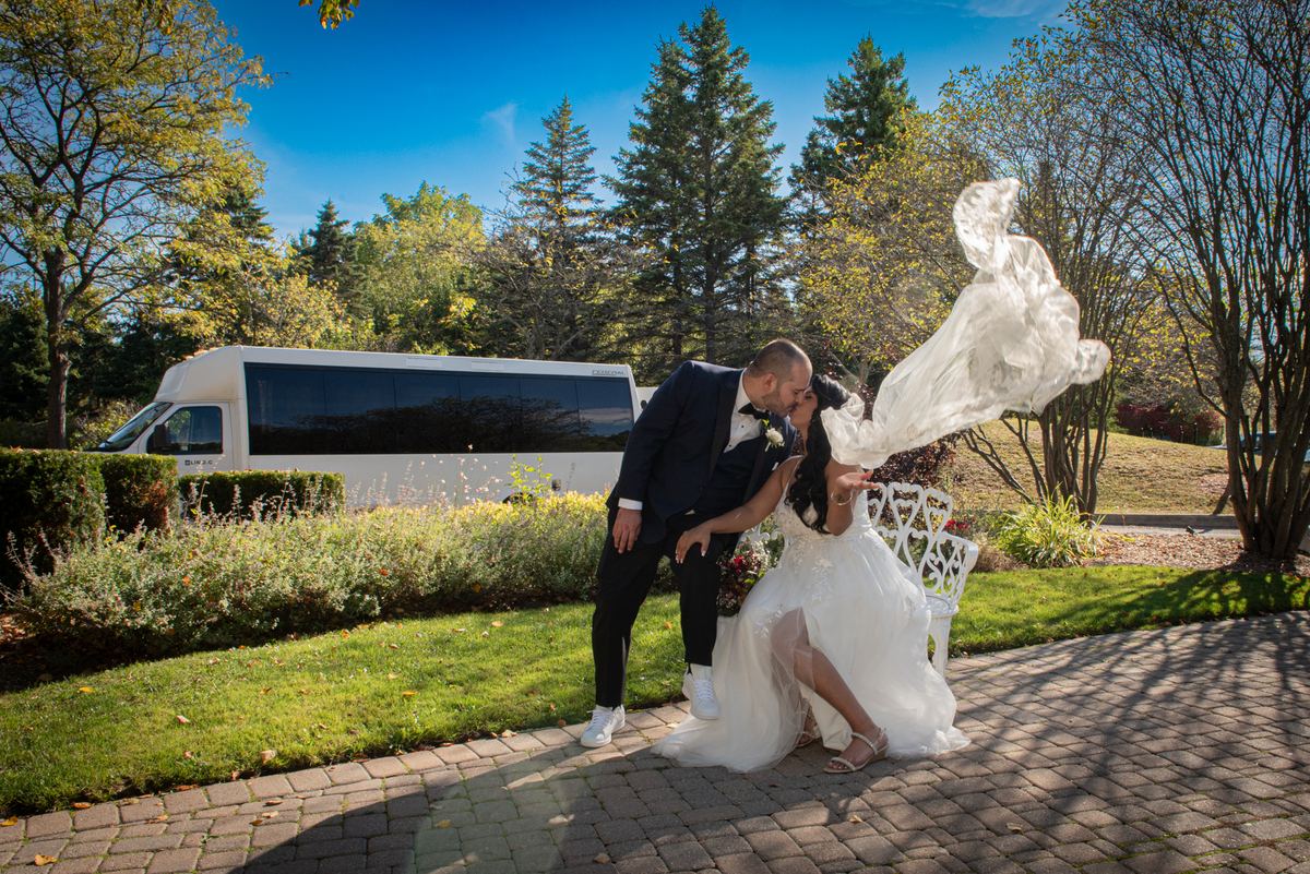 A bride and groom kiss on a garden bench as sunlight filters through autumn foliage, casting shadows on the brick path.