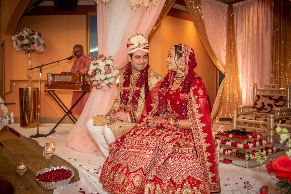 Bride and groom sit on a decorated stage, smiling at each other amid floral arrangements and pink drapery.