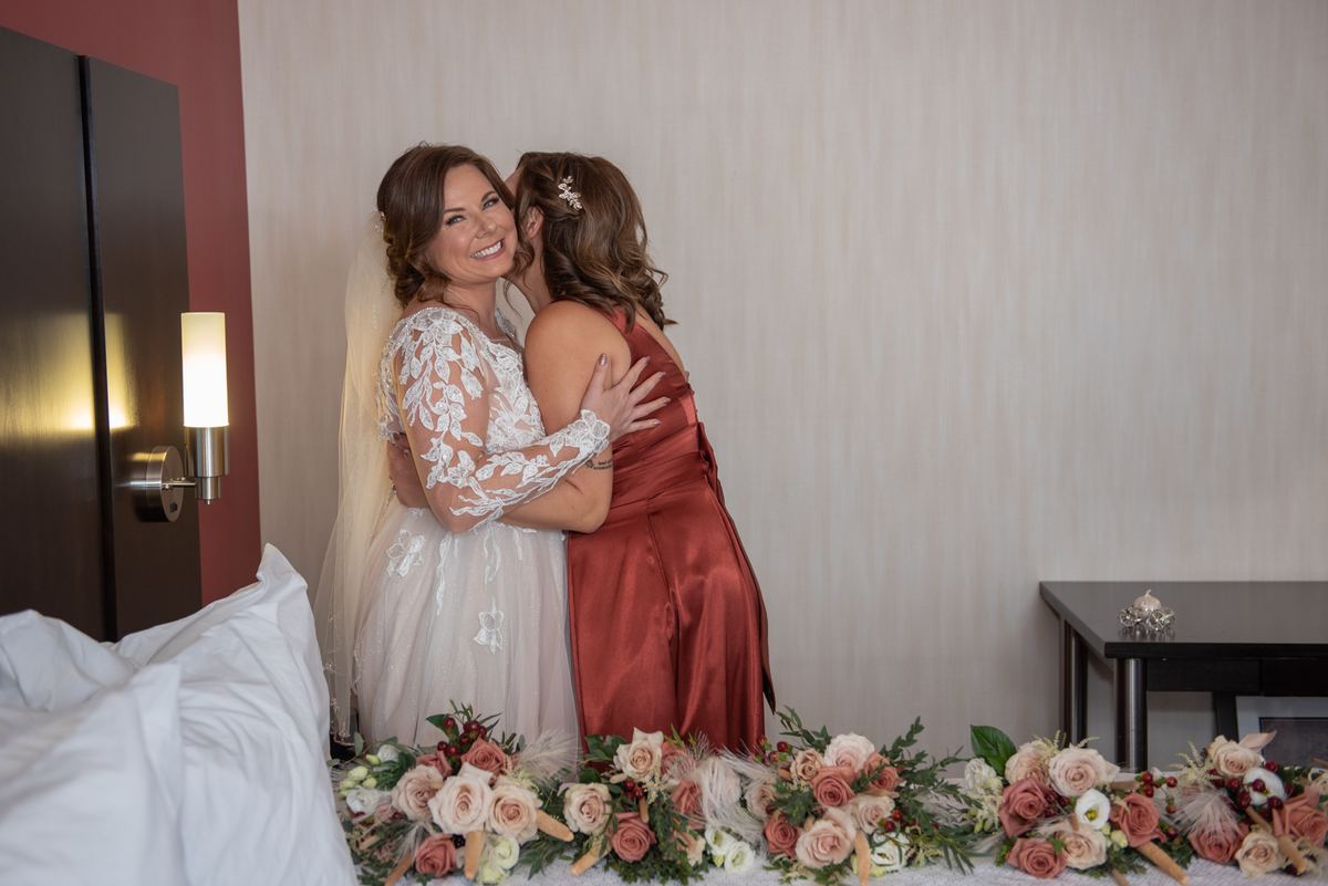 A bride in a white lace gown embraces a family member warmly, surrounded by floral arrangements and soft lighting.
