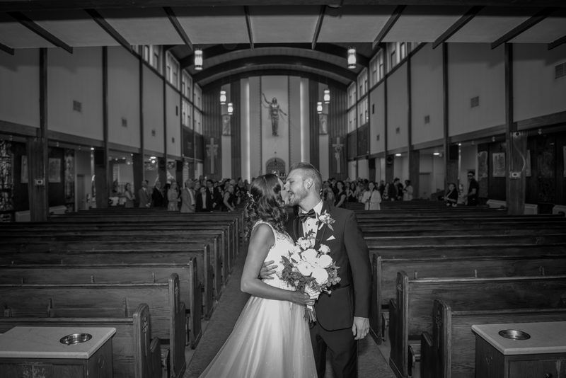 A bride and groom kiss down the aisle of a church surrounded by wooden pews and guests in soft ambient light.