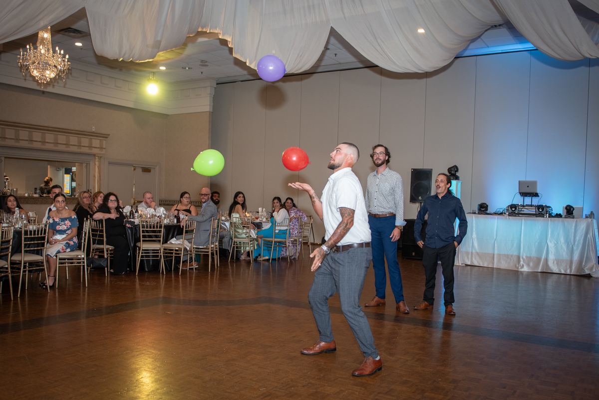 A man in a white shirt juggling red, green, and purple balloons entertains guests at a banquet hall tabled event.
