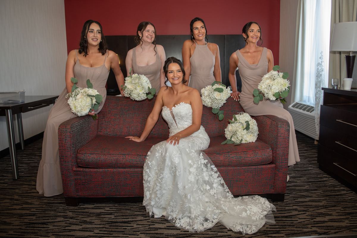 A bride in white sits with four smiling bridesmaids in beige dresses holding hydrangeas on a red couch.