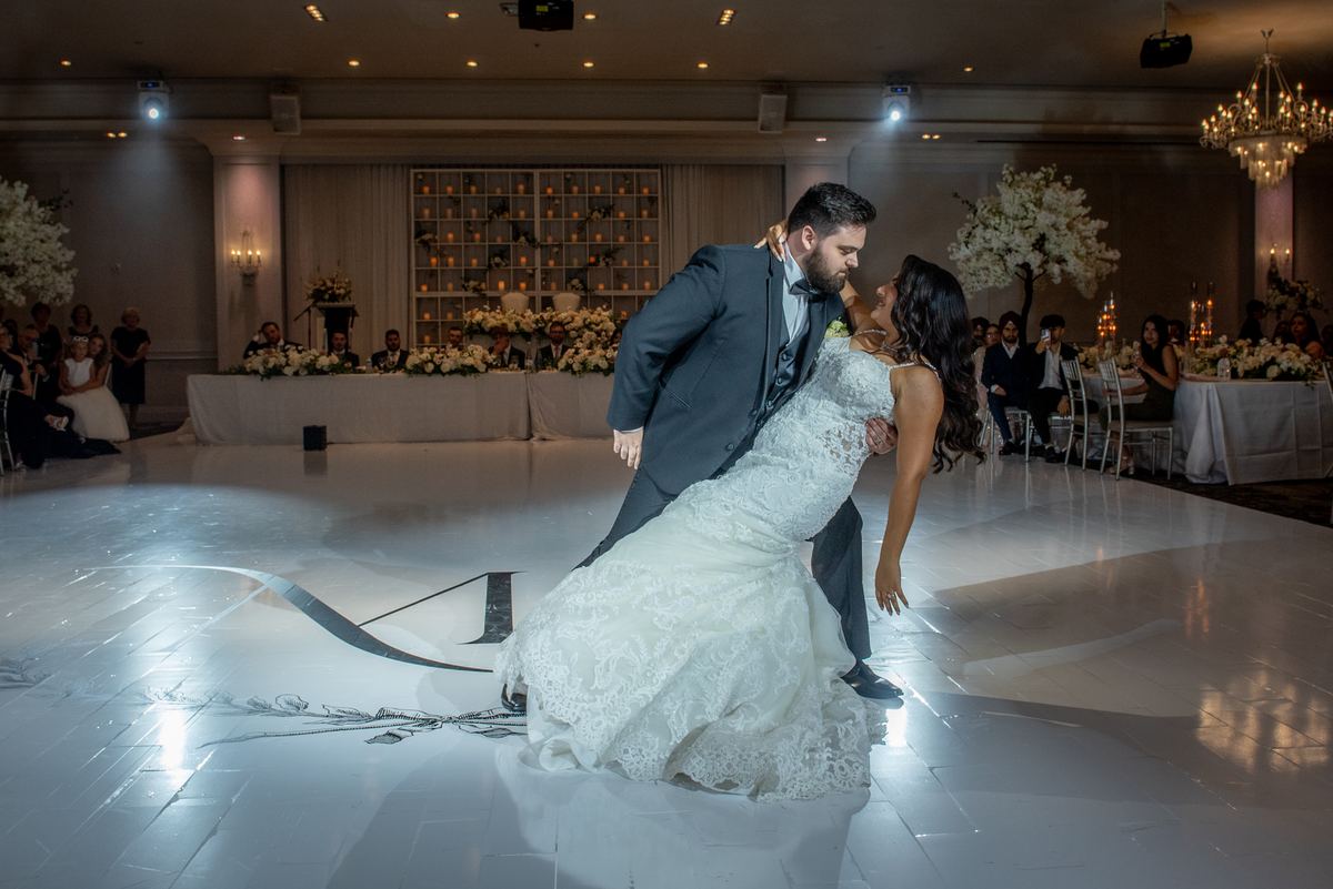 A bride and groom dance on a reflective floor, surrounded by guests at decorated tables in an elegant hall.