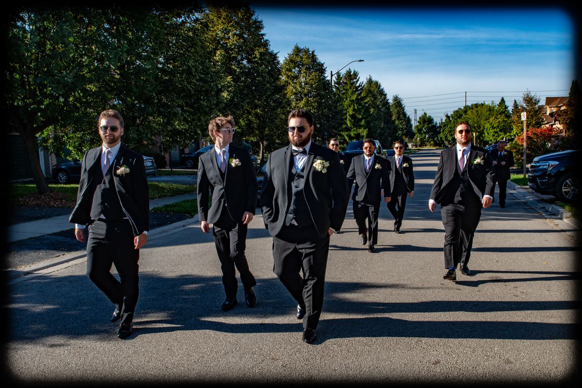 A groom and his groomsmen in black tuxedos walk down a sunlit street, shadows stretching behind them.