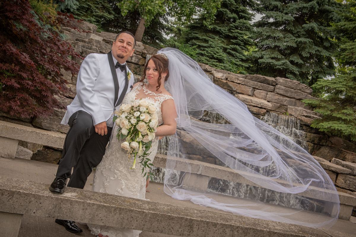 A bride and groom pose for a portrait on stone steps beside a small waterfall, surrounded by lush greenery.